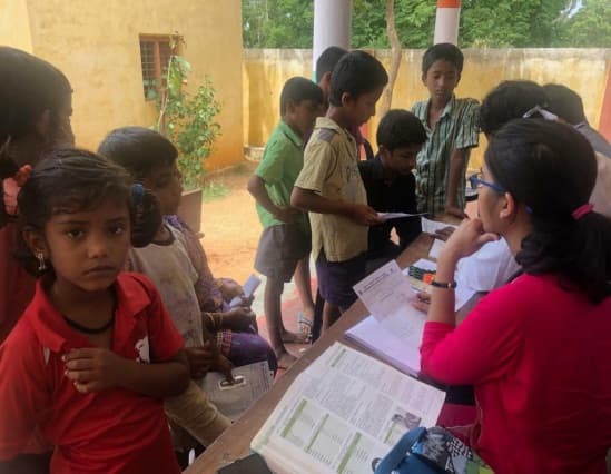 Children in a school courtyard receiving workbooks from Foundation volunteers seated at a table.